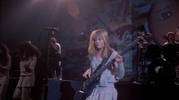 Movie still from “Stop Making Sense” (1984), directed by Jonathan Demme – A woman in a white dress holding a guitar; Wide shot, Low angle