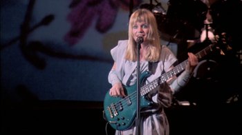 Movie still from “Stop Making Sense” (1984), directed by Jonathan Demme – A woman holding a guitar while standing in front of a microphone; Medium shot, Low angle