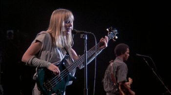 Movie still from “Stop Making Sense” (1984), directed by Jonathan Demme – A woman holding a bass guitar while standing next to a microphone; Medium shot, Low angle