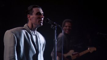 Movie still from “Stop Making Sense” (1984), directed by Jonathan Demme – A man standing on a stage holding a microphone; Medium shot, Low angle