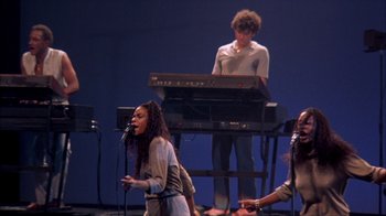 Movie still from “Stop Making Sense” (1984), directed by Jonathan Demme – A group of people on stage singing and playing keyboards; Wide shot, High angle