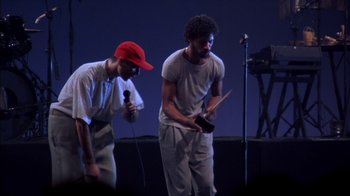 Movie still from “Stop Making Sense” (1984), directed by Jonathan Demme – A couple of men standing next to each other on a stage with microphones; Medium shot, Low angle