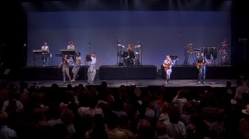 Movie still from “Stop Making Sense” (1984), directed by Jonathan Demme – A group of people on a stage with a crowd watching; Extreme Wide shot, High angle