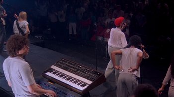 Movie still from “Stop Making Sense” (1984), directed by Jonathan Demme – A group of people standing around a keyboard on a stage; Medium shot, High angle