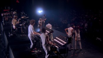 Movie still from “Stop Making Sense” (1984), directed by Jonathan Demme – A group of people playing musical instruments on a stage; Wide shot, High angle