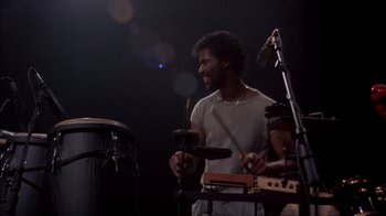Movie still from “Stop Making Sense” (1984), directed by Jonathan Demme – A man is playing drums in a dark room; Medium shot, Low angle