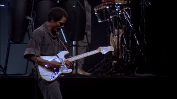 Movie still from “Stop Making Sense” (1984), directed by Jonathan Demme – A man is playing a white guitar on stage; Medium shot, Low angle