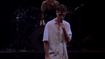 Movie still from “Stop Making Sense” (1984), directed by Jonathan Demme – A man standing on a stage holding a microphone; Medium shot, High angle