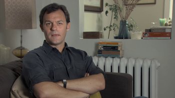 Movie still from “Stories We Tell” (2012), directed by Sarah Polley – A man sitting in a chair with his arms crossed; Close Up shot, Low angle