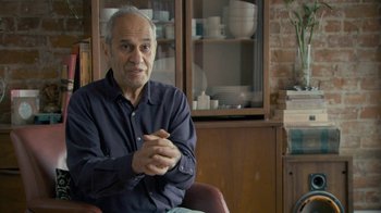 Movie still from “Stories We Tell” (2012), directed by Sarah Polley – An older man sitting in a chair in front of a cabinet; Medium shot, Over the shoulder angle