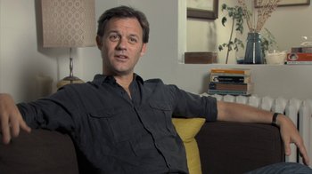 Movie still from “Stories We Tell” (2012), directed by Sarah Polley – A man sitting on top of a couch in a living room; Medium shot, Low angle