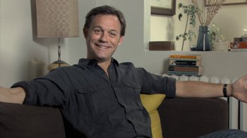 Movie still from “Stories We Tell” (2012), directed by Sarah Polley – A man sitting on a couch holding a book; Medium shot, Low angle