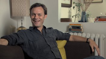 Movie still from “Stories We Tell” (2012), directed by Sarah Polley – A man sitting on a couch smiling for the camera; Medium shot, Low angle