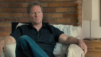 Movie still from “Stories We Tell” (2012), directed by Sarah Polley – A man sitting on top of a couch in a living room; Close Up shot, Over the shoulder angle