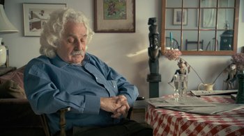 Movie still from “Stories We Tell” (2012), directed by Sarah Polley – An older man sitting at a table with a glass of water; Medium shot, Over the shoulder angle