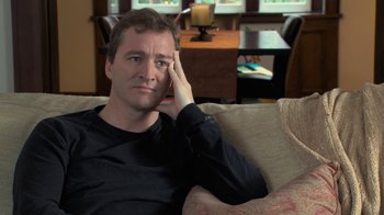 Movie still from “Stories We Tell” (2012), directed by Sarah Polley – A man sitting on top of a couch in a living room; Close Up shot, Over the shoulder angle