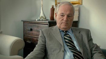 Movie still from “Stories We Tell” (2012), directed by Sarah Polley – An older man wearing a suit and tie sitting in front of a dresser; Close Up shot, Over the shoulder angle