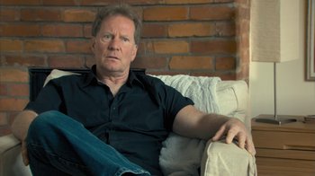 Movie still from “Stories We Tell” (2012), directed by Sarah Polley – A man sitting on top of a couch in a living room; Close Up shot, Over the shoulder angle