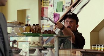 Movie still from “Stranger Than Fiction” (2006), directed by Marc Forster – A man holding a knife in front of a display of cakes; Medium shot, Over the shoulder angle