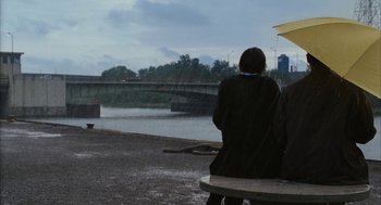 Movie still from “Stranger Than Fiction” (2006), directed by Marc Forster – Two people sitting on a bench in the rain with an umbrella; Wide shot, Over the shoulder angle