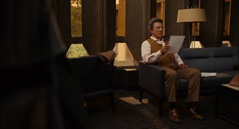 Movie still from “Stranger Than Fiction” (2006), directed by Marc Forster – An older man sitting on a chair reading a paper; Medium shot, Over the shoulder angle