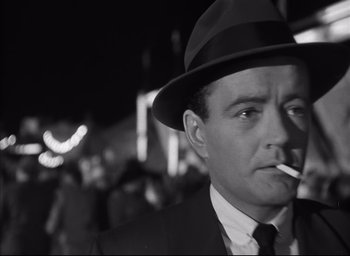 Movie still from “Strangers on a Train” (1951), directed by Alfred Hitchcock – A black and white photo of a man in a suit and hat smoking a cigarette; Close Up shot, Low angle