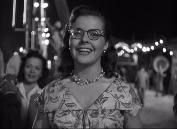 Movie still from “Strangers on a Train” (1951), directed by Alfred Hitchcock – A woman wearing glasses standing in front of a crowd of onlookers; Medium shot, Low angle