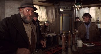 Movie still from “Straw Dogs” (1971), directed by Sam Peckinpah – An old man in a hat is sitting at a bar; Medium shot, Low angle