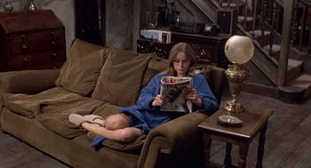Movie still from “Straw Dogs” (1971), directed by Sam Peckinpah – A woman sitting on top of a brown couch reading a magazine; Wide shot, High angle