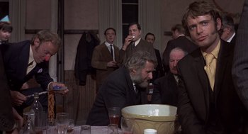 Movie still from “Straw Dogs” (1971), directed by Sam Peckinpah – A group of people sitting at a table with drinks; Medium shot, Over the shoulder angle