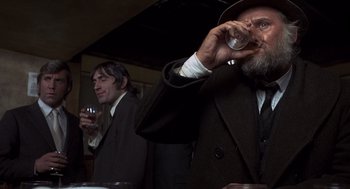 Movie still from “Straw Dogs” (1971), directed by Sam Peckinpah – A man in a suit and a hat is drinking from a wine glass; Close Up shot, Over the shoulder angle