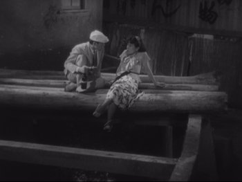 Movie still from “Stray Dog” (1949), directed by Akira Kurosawa – An old photo of a man and a woman sitting on a bench; Wide shot, High angle