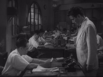 Movie still from “Stray Dog” (1949), directed by Akira Kurosawa – A man standing in front of a table with papers on top of it; Medium shot, Low angle