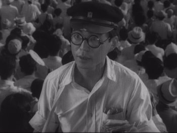 Movie still from “Stray Dog” (1949), directed by Akira Kurosawa – A man wearing a hat and glasses standing in a crowd of people; Close Up shot, High angle