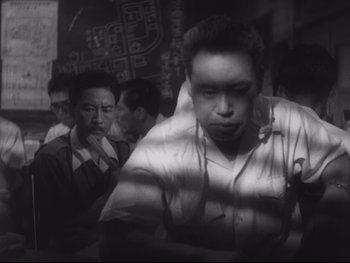 Movie still from “Stray Dog” (1949), directed by Akira Kurosawa – A black - and - white photo of a man in a crowd; Close Up shot, Low angle