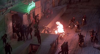 Movie still from “Streets of Fire” (1984), directed by Walter Hill – A group of people standing next to a motorcycle on fire; Wide shot, High angle