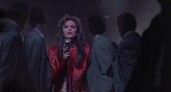 Movie still from “Streets of Fire” (1984), directed by Walter Hill – A woman holding a microphone while standing next to a group of people; Medium shot, Over the shoulder angle