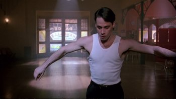 Movie still from “Strictly Ballroom” (1992), directed by Baz Luhrmann – A man in a white tank top and black pants is in a darkened room; Medium shot, Low angle