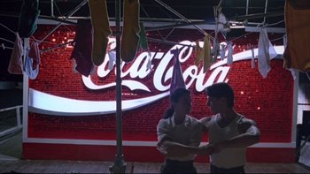 Movie still from “Strictly Ballroom” (1992), directed by Baz Luhrmann – Two men standing next to each other in front of coca - cola sign; Wide shot, Low angle