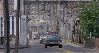 Movie still from “Striking Distance” (1993), directed by Rowdy Herrington – A police car driving down a street next to a bridge; Extreme Wide shot, High angle