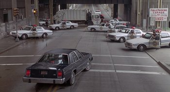 Movie still from “Striking Distance” (1993), directed by Rowdy Herrington – A black car driving down a street next to police cars; Extreme Wide shot, High angle