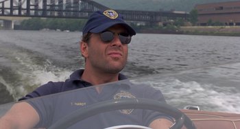 Movie still from “Striking Distance” (1993), directed by Rowdy Herrington – A man wearing a hat driving a boat on a river; Close Up shot, Low angle