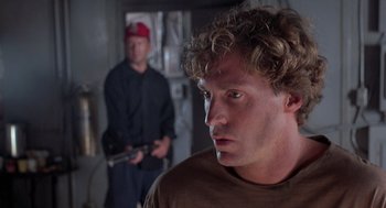Movie still from “Striking Distance” (1993), directed by Rowdy Herrington – A man in a brown t - shirt is looking at the camera while another man stands behind him; Close Up shot, Over the shoulder angle