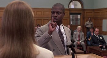 Movie still from “Striking Distance” (1993), directed by Rowdy Herrington – A man in a gray suit is talking to a woman in front of a microphone; Close Up shot, Over the shoulder angle