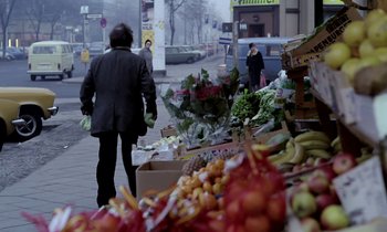 Movie still from “Stroszek” (1977), directed by Werner Herzog – A woman walking down a street next to a fruit and vegetable stand; Wide shot, Over the shoulder angle
