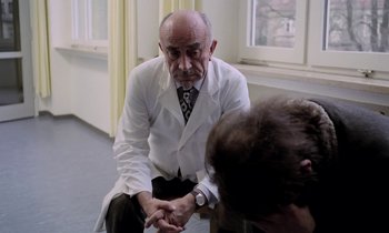 Movie still from “Stroszek” (1977), directed by Werner Herzog – An older man in a white lab coat sits in front of another man; Medium shot, Over the shoulder angle
