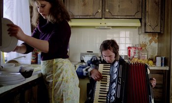 Movie still from “Stroszek” (1977), directed by Werner Herzog – A woman standing next to a man playing an accordion; Medium shot, Low angle