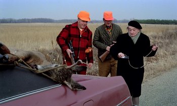 Movie still from “Stroszek” (1977), directed by Werner Herzog – A group of people standing next to a car with a bird on the hood; Medium shot, Over the shoulder angle