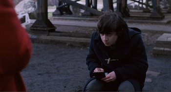 Movie still from “Submarine” (2010), directed by Richard Ayoade – A young man sitting on the ground looking at his cell phone; Medium shot, High angle