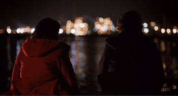 Movie still from “Submarine” (2010), directed by Richard Ayoade – A couple of people standing next to a body of water at night; Medium shot, Over the shoulder angle